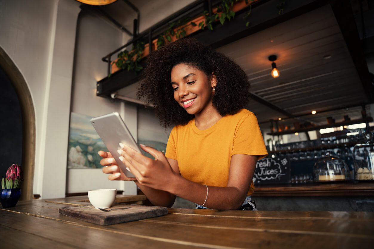 Low angle shot of beautiful African American female holding digital tablet sitting at funky tearoom with hot latte Woman reading tablet while in a café