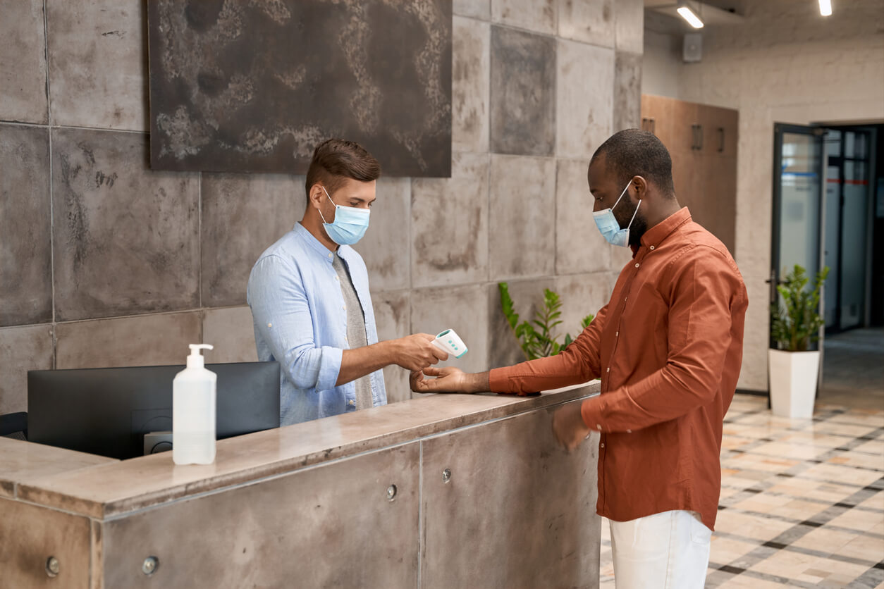 Young male receptionist wearing medical mask standing at reception in the modern office and measuring body temperature of man with infrared thermometer